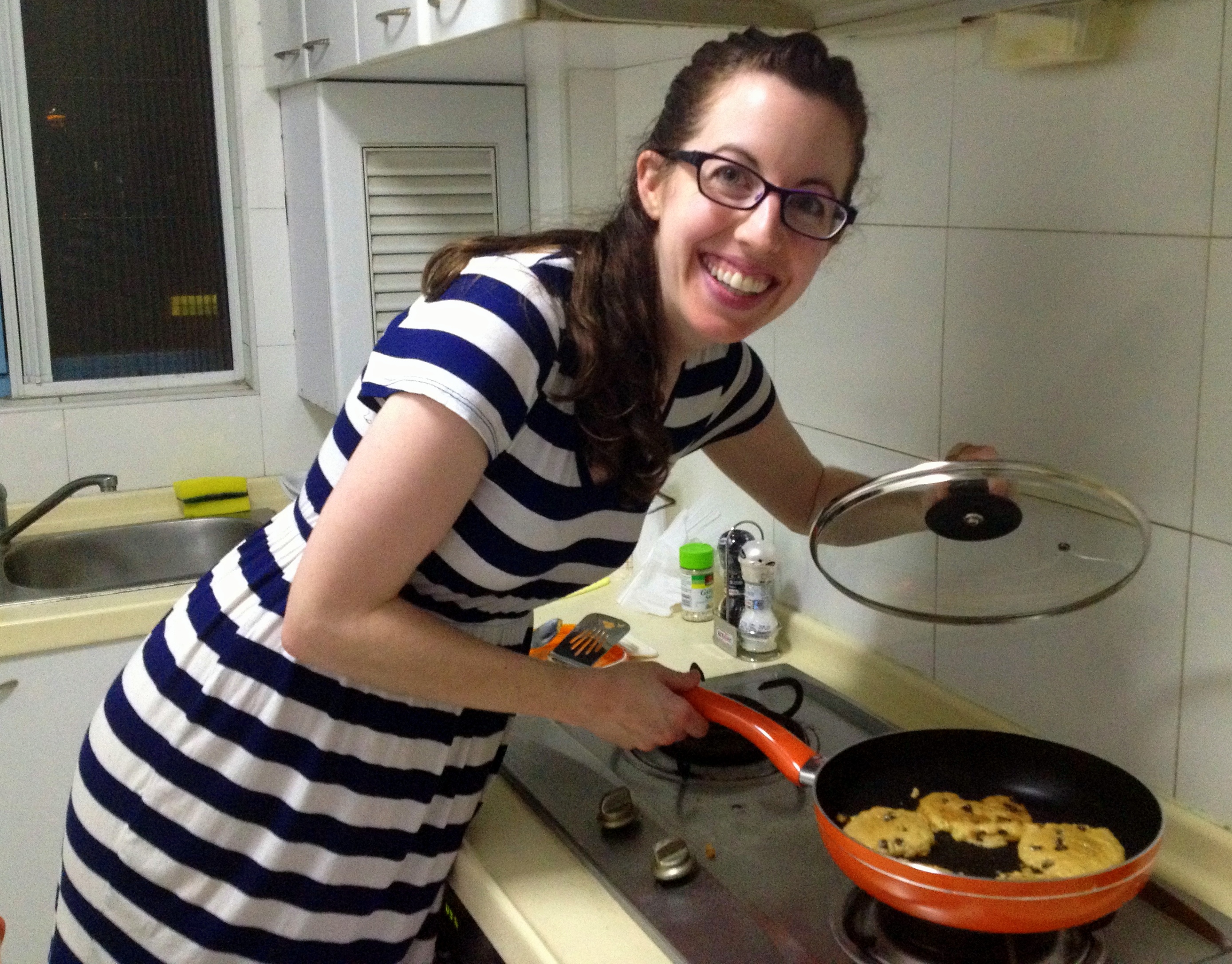 A woman leans over a stove which has cookies in a skillet. She smiles. 