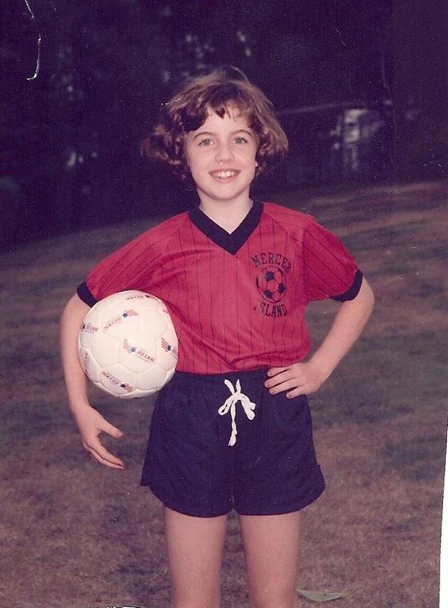 A young girl, aged at approximately 7 years old, wears a red and black soccer uniform and smiles at the camera while holding the ball in one hand.